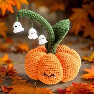 Puede incluir: Una calabaza naranja de crochet con un tallo verde y tres flores blancas en forma de fantasma. La calabaza tiene una cara sonriente linda.