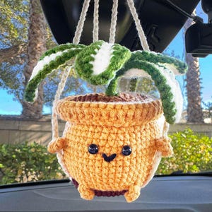 May include: A handmade crochet hanging planter with a smiling face, holding a green and white plant. The planter is yellow and brown, with a whimsical design. The plant is green and white. The planter is hanging from a white string.