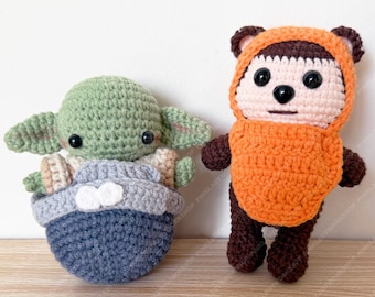May include: Two handmade crochet figures: a green alien-like character in a blue and grey carrier, and a brown bear-like creature in an orange hooded outfit. Both figures have black button eyes, set against a white background.