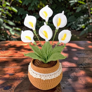 Puede incluir: Un arreglo de lirios de cala hechos a mano con ganchillo en una maceta marrón con un borde de encaje crema. Las flores blancas tienen centros amarillos y tallos y hojas verdes. La maceta está llena de hilo marrón, creando una exhibición floral decorativa.