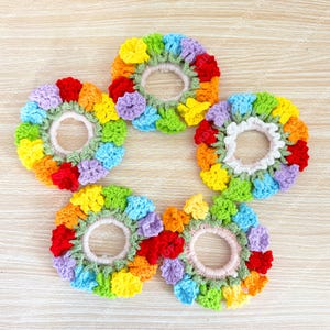 Puede incluir: Cinco coleteros circulares con un diseño de ganchillo de pequeñas flores multicolores en tonos rojo, naranja, amarillo, verde, azul y morado. Cada coletero tiene una base elástica de color claro y hojas tejidas a ganchillo en verde.