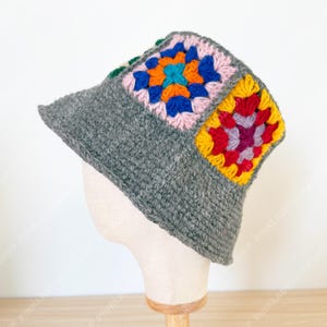 May include: A gray crochet bucket hat with colorful granny square patches. The squares feature vibrant hues of pink, blue, orange, yellow, red, and purple. The hat is displayed on a mannequin head.