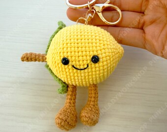 May include: A handmade crochet lemon keychain with a smiling face, black eyes, and brown arms and legs. The yellow lemon has a green leaf and is attached to a gold-colored keyring and clasp.