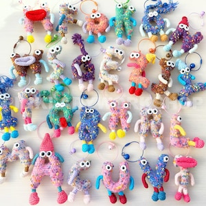 May include: A collection of colorful, handmade crochet keychains in various shapes, including letters and abstract figures. Each keychain features googly eyes and small limbs, with a metal ring for attachment. The keychains are in a variety of colors.