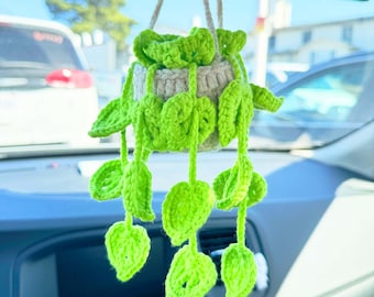 May include: A crocheted green plant hanging from a rearview mirror in a car. The plant has a white pot and green leaves.