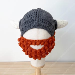 May include: A handmade crochet Viking hat. The hat features a grey helmet, white horns, and an orange beard. The hat is designed for a child and is displayed on a mannequin head.