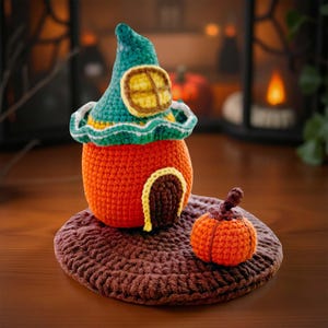 May include: A handmade crochet Halloween decoration featuring an orange pumpkin-shaped house with a teal witch hat and a small pumpkin. The house has a brown door and a yellow window. The items sit on a brown crocheted mat.