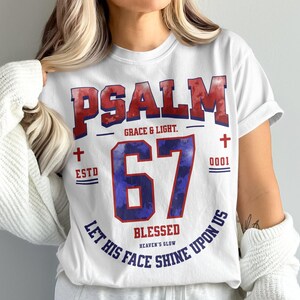 Peut inclure: T-shirt blanc avec l'inscription "PSALM 67" en rouge et bleu. Le t-shirt comprend les phrases "Grace & Light", "Blessed" et "Let His Face Shine Upon Us". Le design comporte des croix et le chiffre "0001".