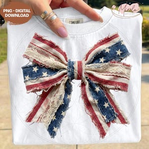 May include: White t-shirt with a large bow design. The bow features red and white striped fabric, blue denim with white stars, and cream-colored lace. The shirt has the word "COMFORT" on the tag.