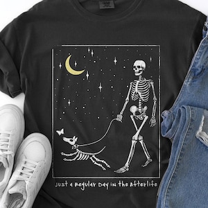 May include: Black t-shirt with a skeleton walking a skeleton dog under a crescent moon and stars. The text "just a regular day in the afterlife" is printed on the shirt. The image also includes a pair of white sneakers and blue jeans.