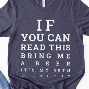 May include: A dark heather navy blue short-sleeve t-shirt featuring white text printed in an eye chart style that reads: IF YOU CAN READ THIS BRING ME A BEER IT'S MY 40TH BIRTHDAY. The shirt has a crew neckline and a standard fit design.