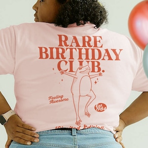 May include: Pink t-shirt with the text "RARE BIRTHDAY CLUB." in red. A cartoon frog with outstretched arms is above the text. The text "Feeling Awesome" is below the frog. The text "Est. 1984" is below the frog's feet.