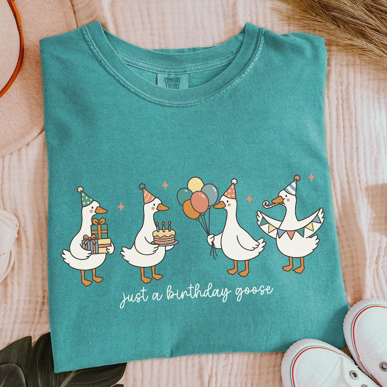 May include: Teal-colored t-shirt featuring a whimsical illustration of four geese wearing party hats. Each goose holds birthday-themed items like gifts, a cake, and balloons. The text "just a birthday goose" is written in cursive below the geese.