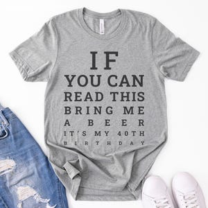 May include: A heather gray short-sleeve t-shirt with black text printed in an eye chart style that reads: IF YOU CAN READ THIS BRING ME A BEER IT'S MY 40TH BIRTHDAY. The shirt features a crew neckline and is displayed alongside blue denim jeans.
