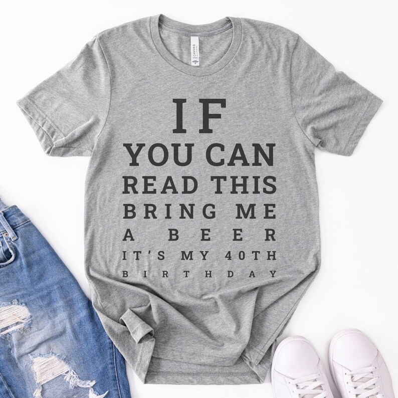 May include: A heather gray short-sleeve t-shirt with black text printed in an eye chart style that reads: IF YOU CAN READ THIS BRING ME A BEER IT'S MY 40TH BIRTHDAY. The shirt features a crew neckline and is displayed alongside blue denim jeans.