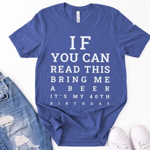 May include: A heather blue short-sleeve t-shirt featuring white text arranged like an eye chart that reads: IF YOU CAN READ THIS BRING ME A BEER IT'S MY 40TH BIRTHDAY. The shirt has a crew neckline and a standard fit, displayed flat with a casual style.