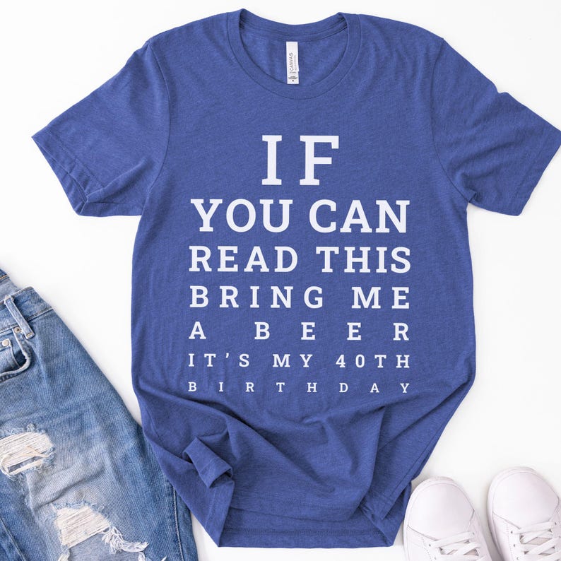 May include: A heather blue short-sleeve t-shirt featuring white text arranged like an eye chart that reads: IF YOU CAN READ THIS BRING ME A BEER IT'S MY 40TH BIRTHDAY. The shirt has a crew neckline and a standard fit, displayed flat with a casual style.