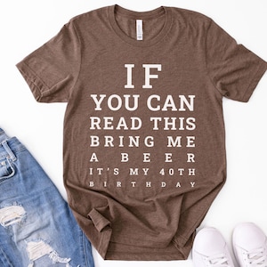 May include: A brown short-sleeved t-shirt featuring white text that reads: IF YOU CAN READ THIS BRING ME A BEER IT'S MY 40TH BIRTHDAY. The shirt is displayed alongside blue denim jeans and white sneakers, showcasing a casual style for a birthday.