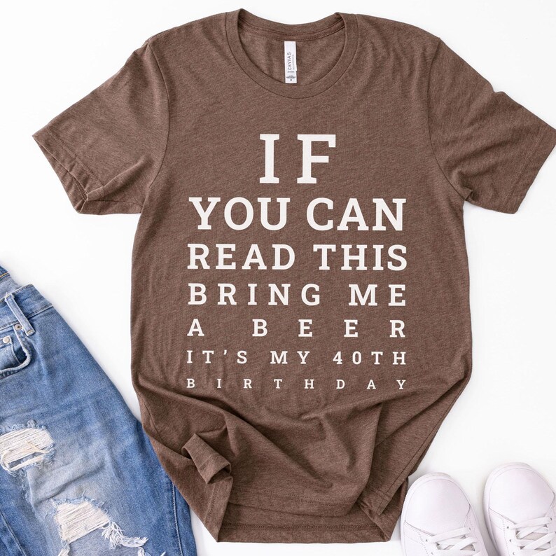 May include: A brown short-sleeved t-shirt featuring white text that reads: IF YOU CAN READ THIS BRING ME A BEER IT'S MY 40TH BIRTHDAY. The shirt is displayed alongside blue denim jeans and white sneakers, showcasing a casual style for a birthday.