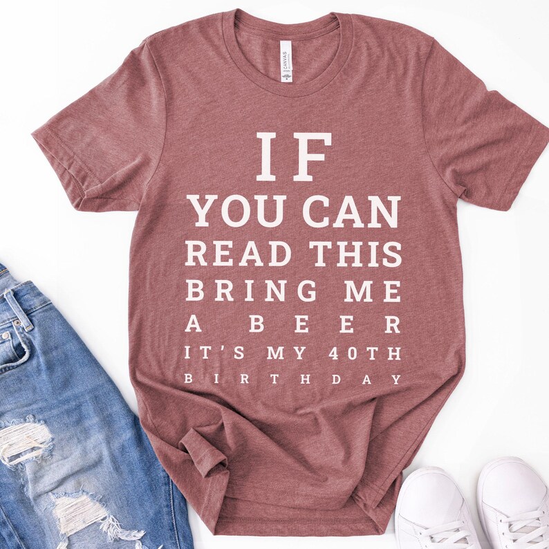 May include: A heathered mauve short-sleeve t-shirt with white text that reads: IF YOU CAN READ THIS BRING ME A BEER IT'S MY 40TH BIRTHDAY. The shirt features a crew neckline and a standard fit, displayed alongside blue denim jeans and white sneakers.