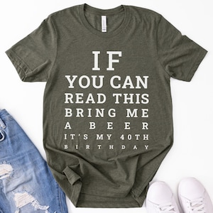 May include: An olive green short-sleeved t-shirt featuring white text printed in an eye chart style that reads: IF YOU CAN READ THIS BRING ME A BEER IT'S MY 40TH BIRTHDAY. The shirt has a crew neckline and a standard fit, displayed flat.