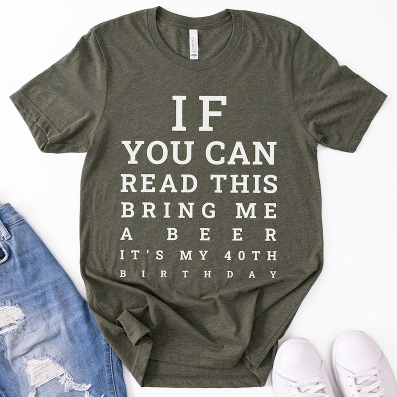May include: An olive green short-sleeved t-shirt featuring white text printed in an eye chart style that reads: IF YOU CAN READ THIS BRING ME A BEER IT'S MY 40TH BIRTHDAY. The shirt has a crew neckline and a standard fit, displayed flat.