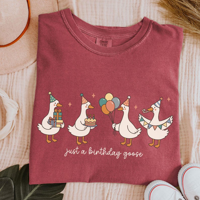 May include: Burgundy t-shirt featuring a whimsical illustration of four geese celebrating a birthday. Each goose wears a party hat and carries gifts, a cake, or balloons. The text "just a birthday goose" is printed below the geese.