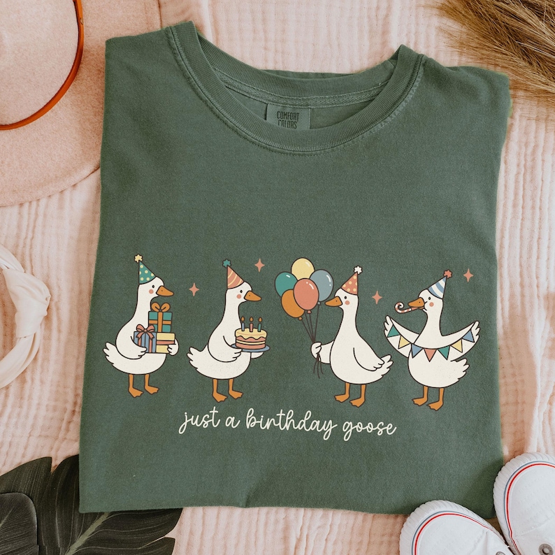 May include: Green t-shirt featuring four cartoon geese celebrating a birthday. Each goose wears a party hat and holds gifts, a cake, balloons, or a banner. The text "just a birthday goose" is below the geese.