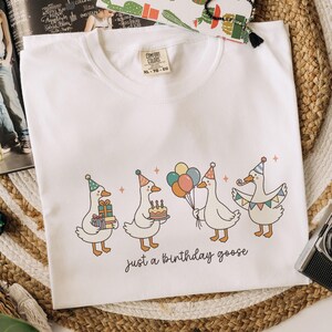 May include: White crew neck t-shirt with a whimsical illustration of four geese celebrating a birthday. Each goose wears a party hat and holds gifts, a cake, or balloons. The text "just a birthday goose" is printed below the geese.