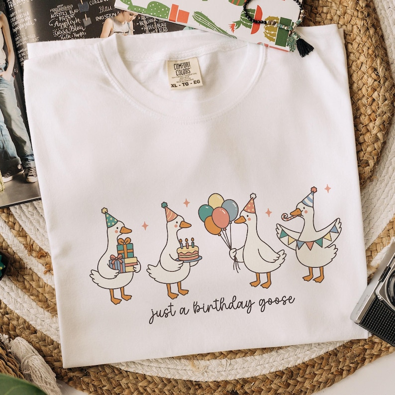 May include: White crew neck t-shirt with a whimsical illustration of four geese celebrating a birthday. Each goose wears a party hat and holds gifts, a cake, or balloons. The text "just a birthday goose" is printed below the geese.
