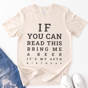 May include: A cream-colored short-sleeve t-shirt featuring black text arranged like an eye chart. The text reads: IF YOU CAN READ THIS BRING ME A BEER IT'S MY 40TH BIRTHDAY. The shirt has a crew neckline and is made of a soft, lightweight fabric.