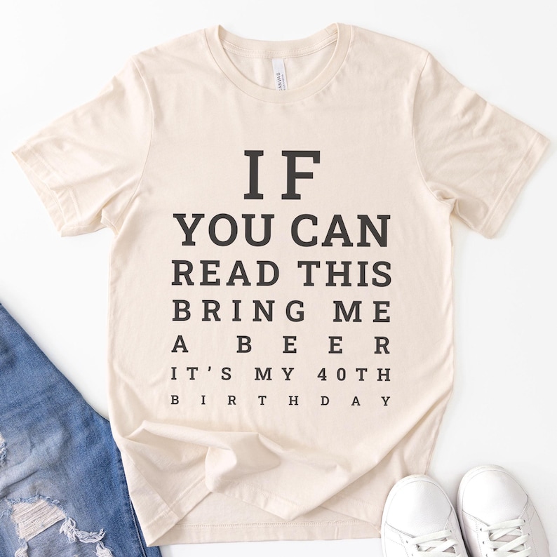 May include: A cream-colored short-sleeve t-shirt featuring black text arranged like an eye chart. The text reads: IF YOU CAN READ THIS BRING ME A BEER IT'S MY 40TH BIRTHDAY. The shirt has a crew neckline and is made of a soft, lightweight fabric.