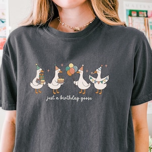 May include: Dark gray t-shirt featuring a whimsical illustration of four geese celebrating a birthday. Each goose wears a party hat and carries gifts, a cake, or balloons. The text "just a birthday goose" is printed below the geese.