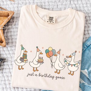 May include: Cream-colored t-shirt featuring a whimsical illustration of four geese celebrating a birthday. Each goose wears a party hat and holds gifts, a cake, balloons, and a banner. The text "just a birthday goose" is below the geese.