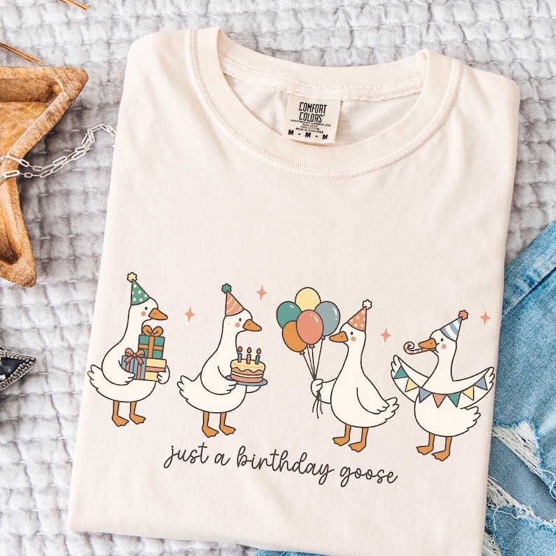 May include: Cream-colored t-shirt featuring a whimsical illustration of four geese celebrating a birthday. Each goose wears a party hat and holds gifts, a cake, balloons, and a banner. The text "just a birthday goose" is below the geese.