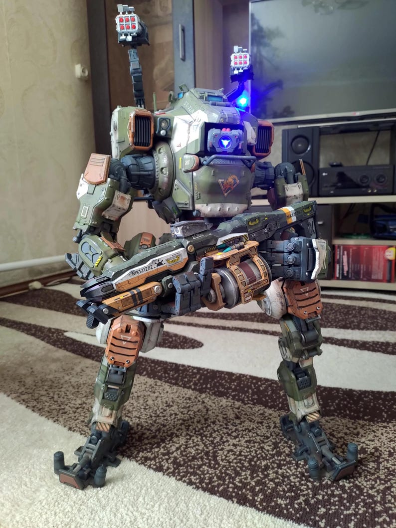 May include: A detailed model of a military-style robot, featuring a color scheme of green, brown, and orange. The robot has multiple weapons and a complex mechanical design. It stands on two legs and has two missile launchers on its shoulders.