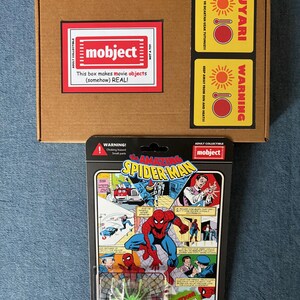 Pu&ograve; includere: Una scatola di cartone marrone con un'etichetta rossa e bianca con la scritta "mobject" e "This box makes movie objects (somehow) REAL!". Sotto, una action figure "The Amazing Spider-Man" in stile vintage in un blister. La confezione include un ragno verde.
