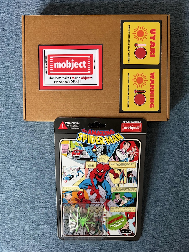 Pu&ograve; includere: Una scatola di cartone marrone con un'etichetta rossa e bianca con la scritta "mobject" e "This box makes movie objects (somehow) REAL!". Sotto, una action figure "The Amazing Spider-Man" in stile vintage in un blister. La confezione include un ragno verde.