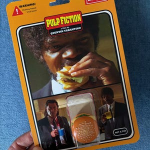 May include: A Pulp Fiction collectible figure in a retro-style orange package. The packaging features images from the film, including Samuel L. Jackson eating a burger. The package also includes the text "WARNING! Choking hazard Small parts."