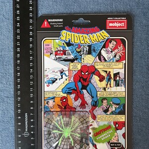 Pu&ograve; includere: Un personaggio d'azione di Spider-Man in una confezione di plastica. La confezione presenta fumetti con Spider-Man in azione. Un giocattolo a forma di ragno verde &egrave; visibile in basso. La confezione include il testo "WARNING! Choking hazard Small parts" e "CAUTION! RADIOACTIVE SPIDER".
