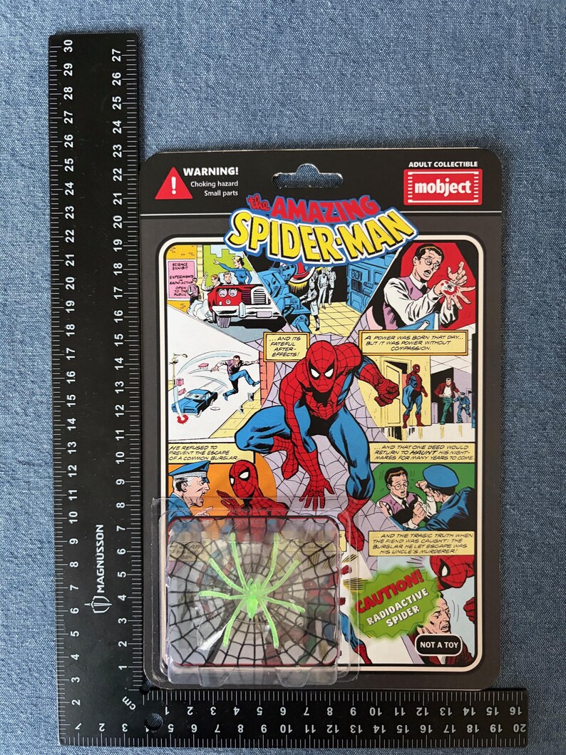 Pu&ograve; includere: Un personaggio d'azione di Spider-Man in una confezione di plastica. La confezione presenta fumetti con Spider-Man in azione. Un giocattolo a forma di ragno verde &egrave; visibile in basso. La confezione include il testo "WARNING! Choking hazard Small parts" e "CAUTION! RADIOACTIVE SPIDER".
