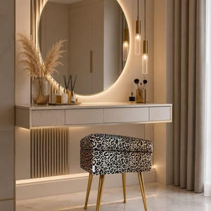 May include: A wall-mounted vanity table with a white finish, four drawers, and gold trim. Above it, a large round backlit mirror. A stool with a black and white leopard print fabric seat and four slender gold metal legs sits in front of the vanity.