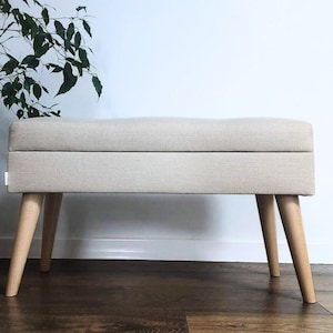 May include: A rectangular storage bench upholstered in a light beige fabric with a smooth, flat top. The bench is supported by four light-coloured wooden legs that taper towards the floor. A small white fabric tag is visible on the side of the bench.