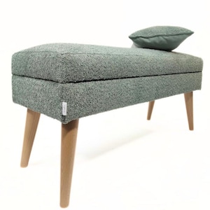 Può includere: Una panca imbottita verde chiaro con gambe in legno e un cuscino abbinato. La panca ha un design trapuntato e una forma rettangolare.