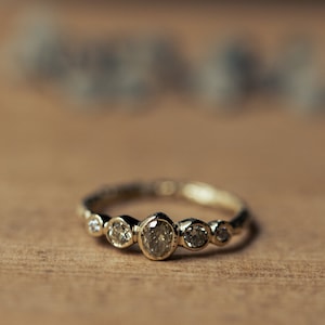 Op de afbeelding: Een gouden ring met een reeks kleine, ronde, heldere edelstenen. De ring heeft een delicate band en de edelstenen zijn in een bezel-stijl gezet. De ring wordt gepresenteerd op een houten oppervlak.