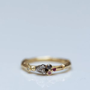 May include: A gold ring with a unique design. The ring features a cluster of gemstones, including a black diamond, a ruby, and a pink sapphire. The band has a textured, organic appearance.
