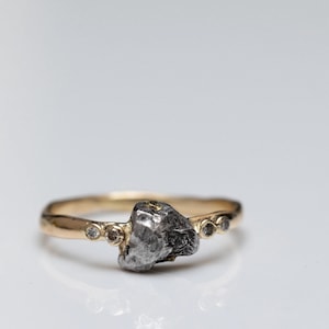 May include: A gold ring featuring a rough, dark gray stone as the centerpiece. The band is a warm gold color, with small, clear stones set on either side of the main stone. The ring is set against a white background.