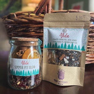 May include: Two glass jars filled with a Yule simmer pot blend. The jar on the left is labeled "Yule Simmer Pot Blend" and the jar on the right is labeled "Yule" with a list of ingredients: cinnamon, orange peel, anise, ginger, cloves, black tea, crushed candy cane. The jars are sitting on a wooden table next to a brown paper bag with a label that says "The Witch's Brew".