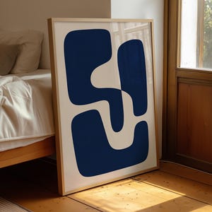 May include: A framed wall art print featuring a large, dark navy blue abstract shape with rounded edges set against a solid off-white background. The artwork is enclosed in a light-toned wooden frame, displaying a bold, fluid, and organic design.
