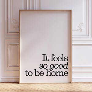 May include: A framed art print with the text "It feels so good to be home" in black lettering. The print has a light wood frame and is leaning against a white wall with decorative molding. The floor is made of wood.